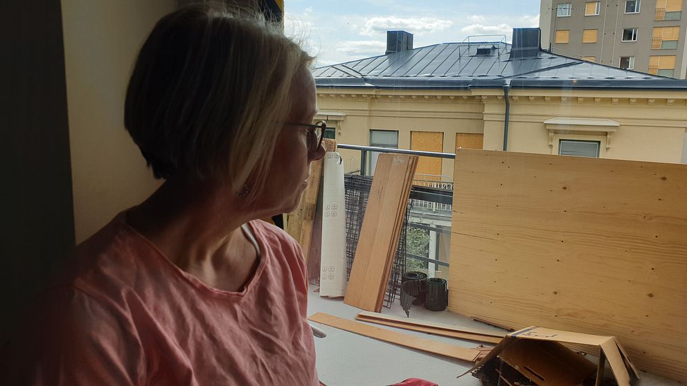 Annika tittar ut på balkongen som är full av bråte och brädor, på glaset sitter gulsvart tejp över sprickorna. I bakgrunden syns två hus med träskivor för många av fönstren.