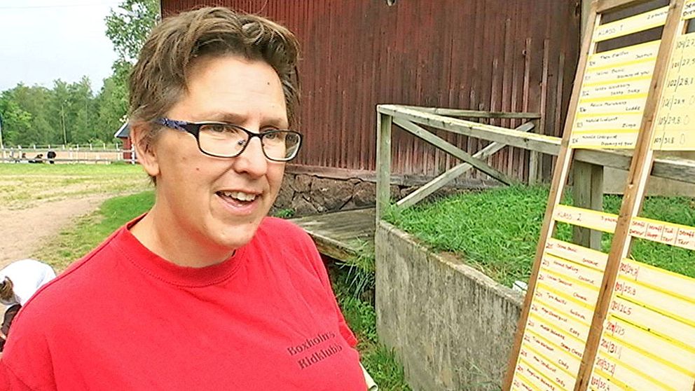 Kvinna med kort hår, glasögon och röd t-shirt framför ett stall