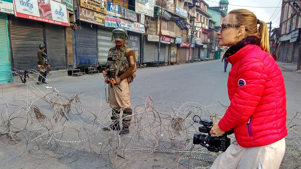 Kvinna i röd jacka står på gata och filmar med videokamera. I bakgrunden syns två soldater med vapen och avspärrning.