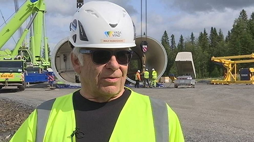 Bild på Rolf Rubensson  klädd i varselkläder och bygghjälm framför arbetare som reser ett vindkraftverk i Åskälen
