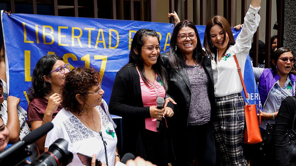 Fem kvinnor står på en liten scen. I mitten är nu frikända Evelyn Hernandez klädd i svar och rosa, med en mikrofon i handen.
