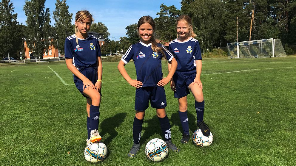 Tre flickor let mot kameran iklädda fotbolls-ställ på en fotbollsplan med träd i bakgrunden.