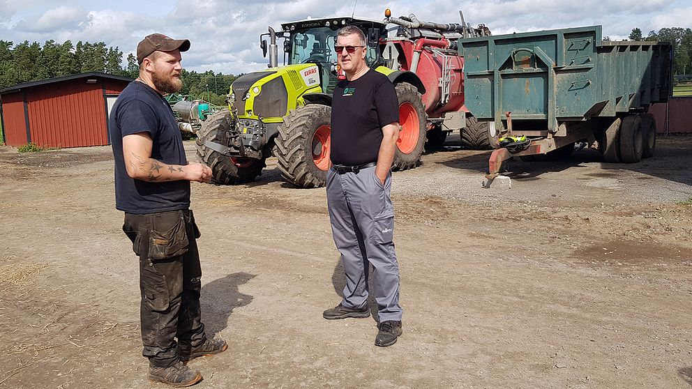 Två män som står på en ladugårdplan framför traktorer.