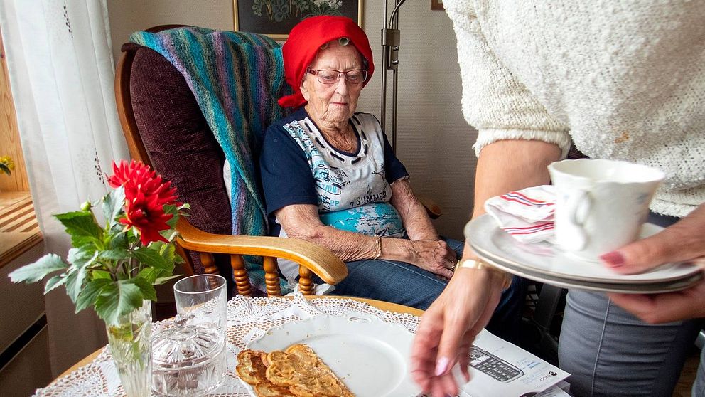 Äldre dam sitter vid ett bord och hemtjänstpersonal plockar bort disk