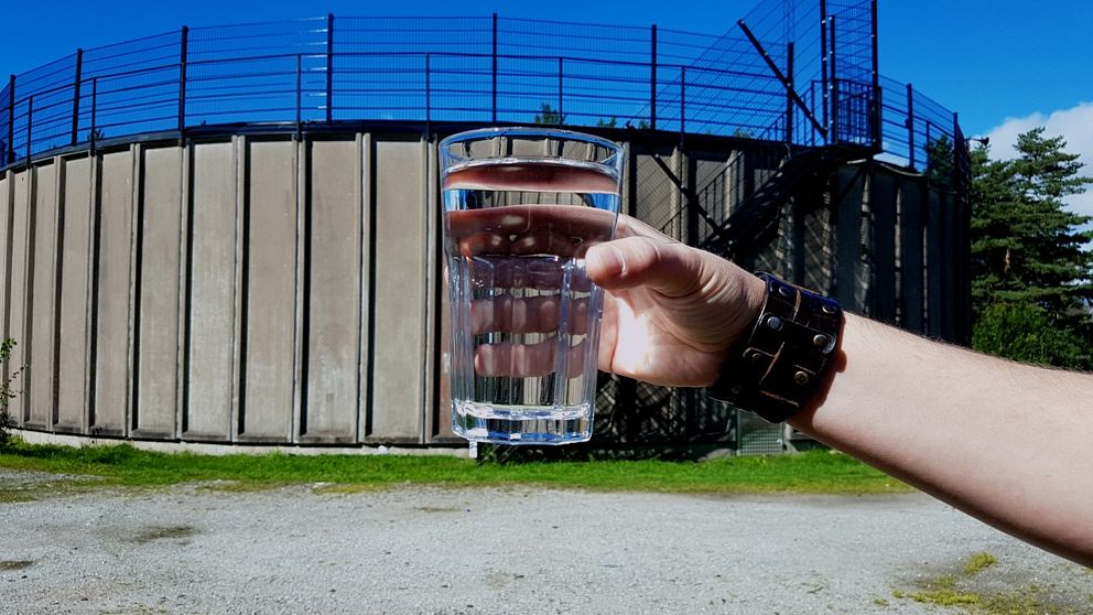 Hand som håller i ett vattenglas i förgrunden. I bakgrunden syns vattenverket i Stenungsund.