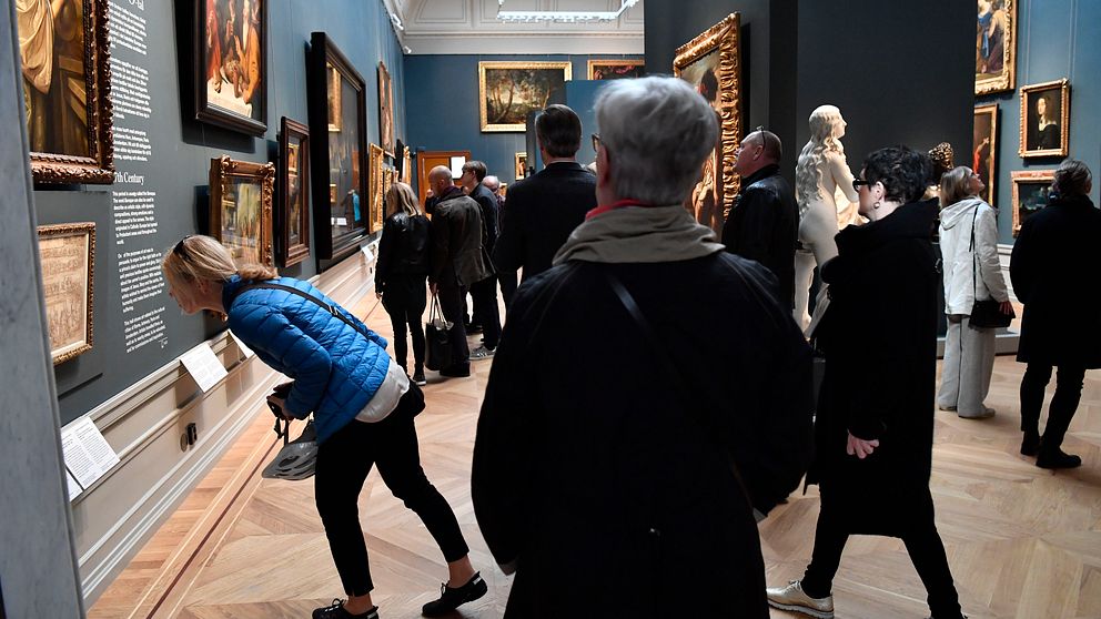 Många besökare tittar på en utställning. En kvinna lutar sig framåt och tittar noga på en tavla.