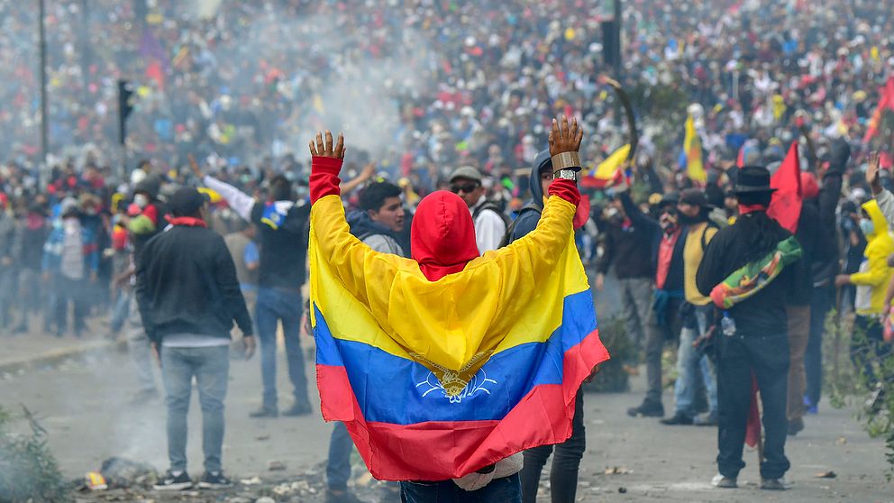 Bild på en protesterande folkmassa med en person som har Ecuadors flagga runt ryggen. Demonstrationerna sker mot president Morenos ekonomiska politik.
