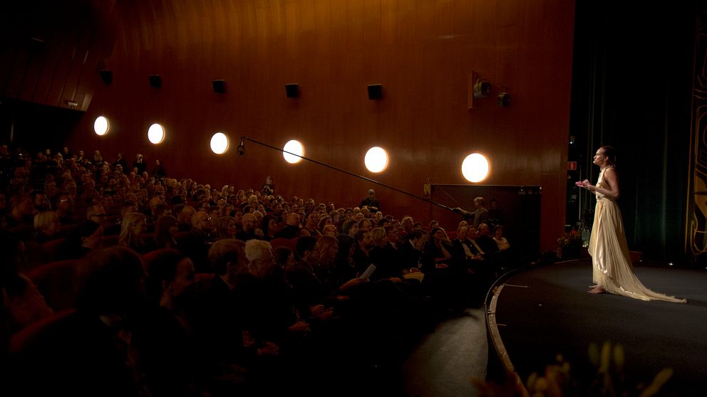 Skådespelaren Bianca Kronlöf står på scen i finklänning inför en publik.
