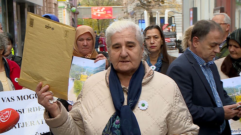 Flera personer står på en gata och protesterar. I mitten en äldre dam som håller upp ett stort kuvert.