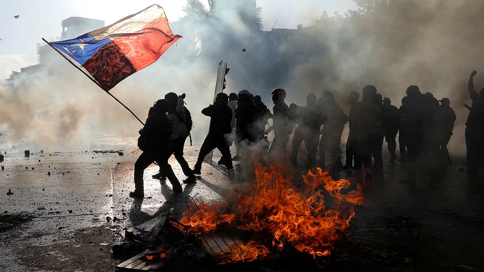 Demonstranter i Chile håller upp en chilensk flaga och står intill bråte som står i lågor.