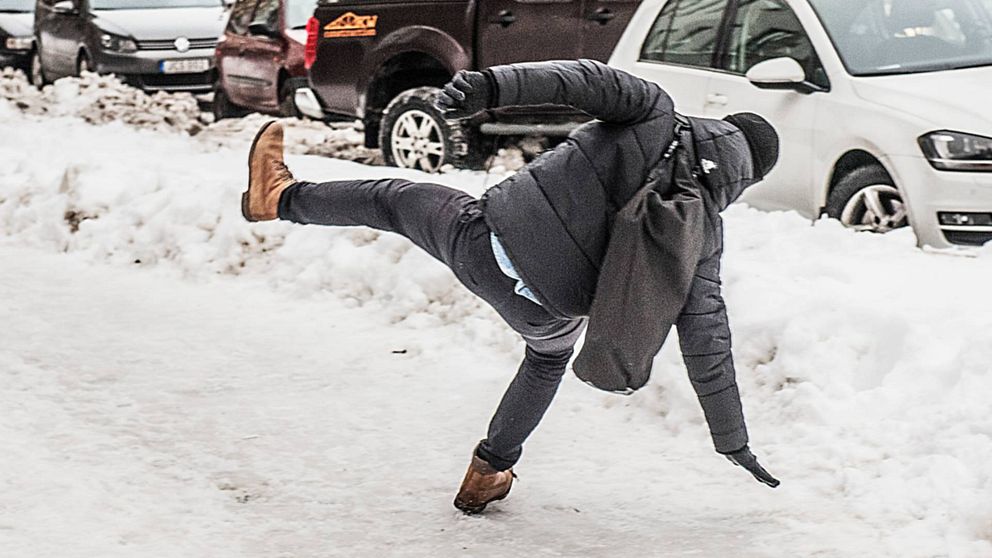 En man iklädd svarta vinterkläder och ryggsäck halkar på en snöbeklädd gata.