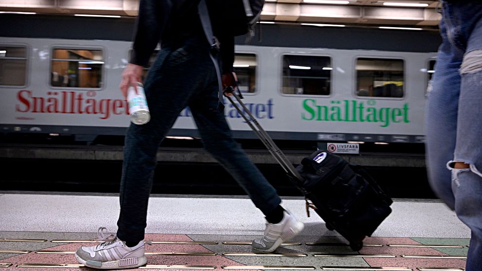 Person går med rullväska på perrong. I bakgrunden ett Snälltåg.