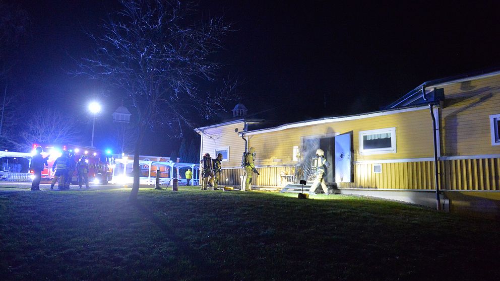 Brandbilar och brandmän står utanför samlingslokal, en gul träbyggnad. Det ryker från dörren.