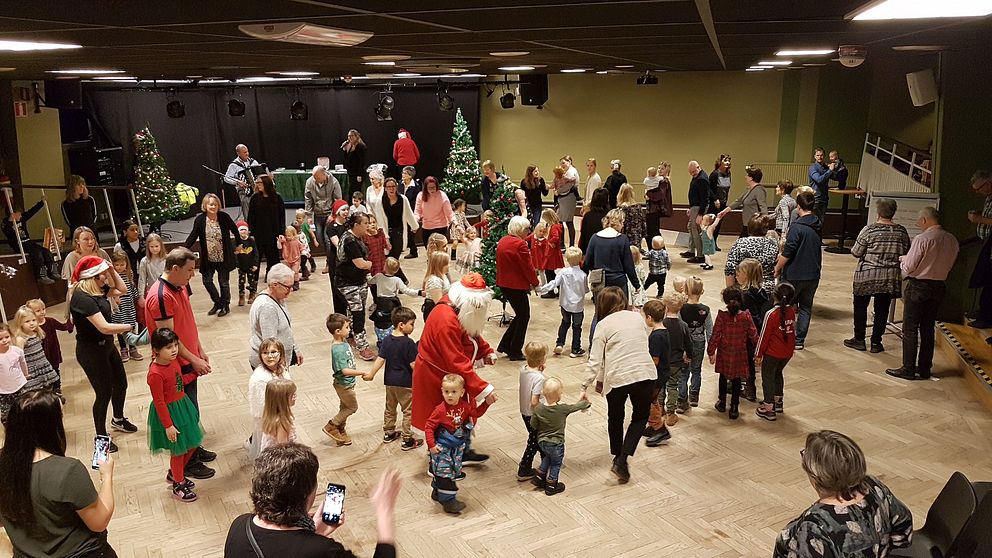 Barn, tomtar och vuxna dansar runt en julgran.