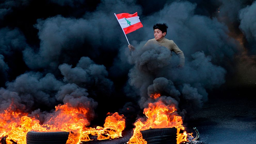 En pojke hoppar över brända bildäck, genom svart rök, med en libanesisk flagga i handen.
