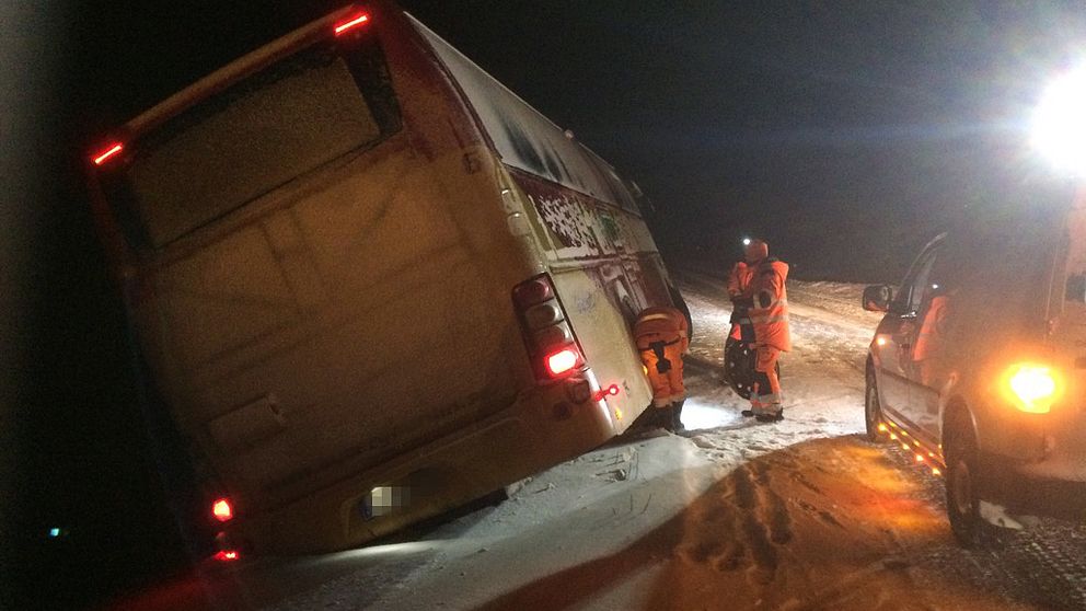 en buss står kraftigt lutande på sidan av snöig väg, räddningspersonal utanför