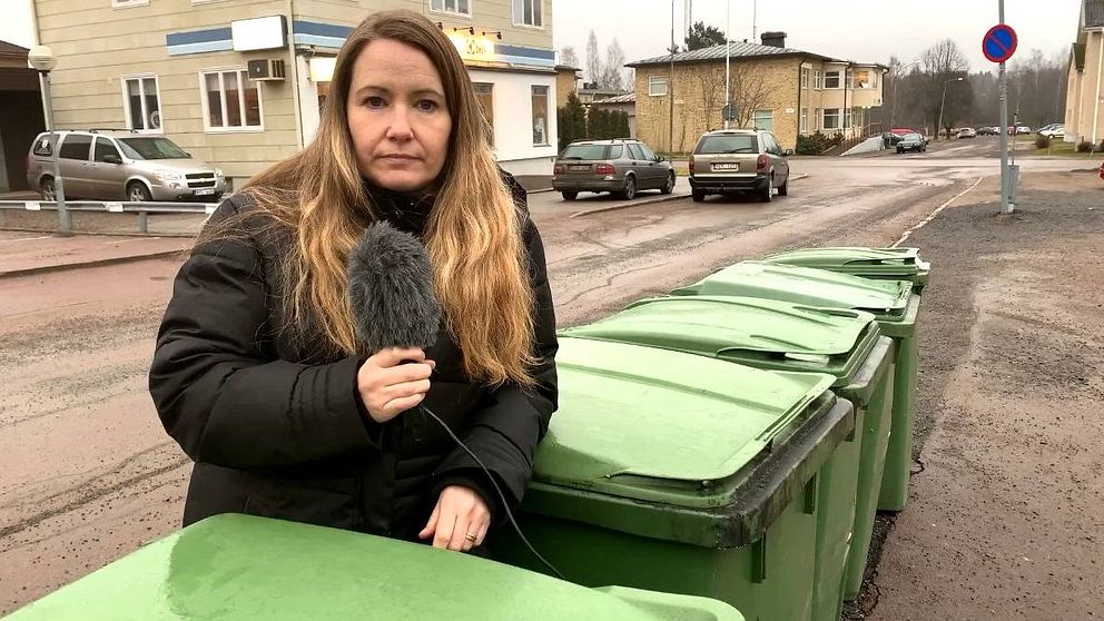 kvinna håller i mick och står framför flera gröna soptunnor.