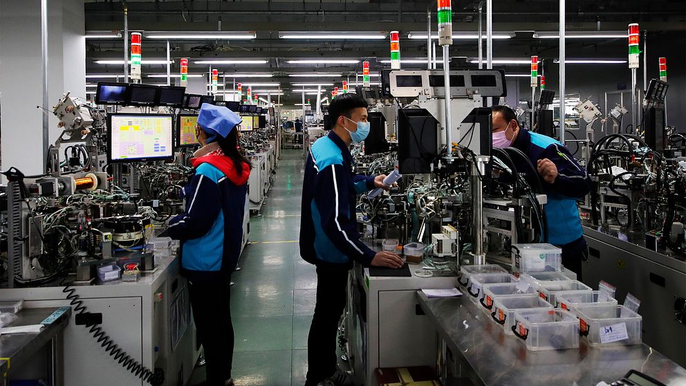 tre fabriksarbetare står framför datorer och jobbar. Alla bär munskydd och marinblåa arbetskläder.
