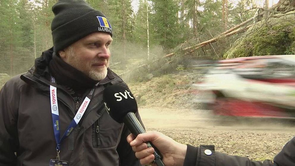 Jason Downey står i skogen, med en mössa som det står Barbados på, medan en rallybil passerar i bakgrunden.