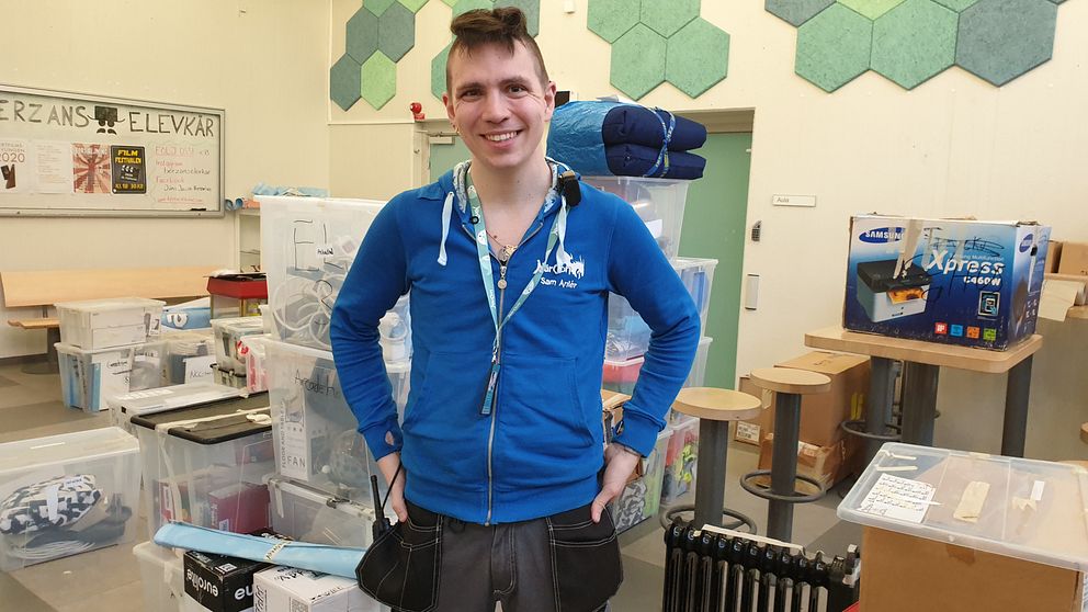 Sam står i blå träningsjacka bland plastlådor med prylar som ska packas upp, med armarna i sidorna.