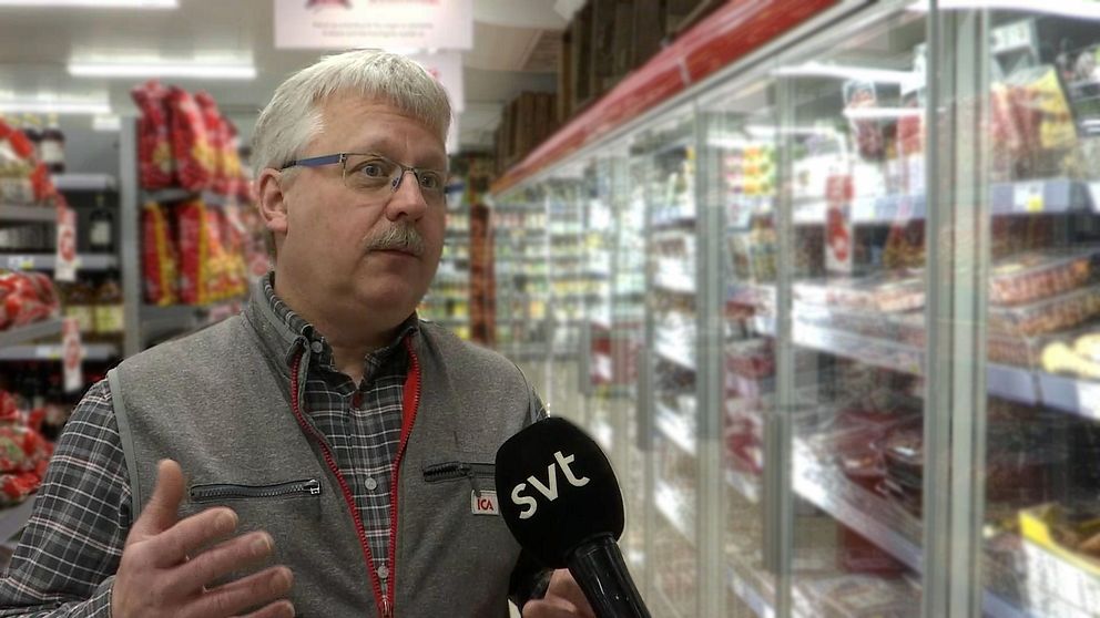 Man med mustasch och glasögon står i en matbutik med en mikrofon framför ansiktet.