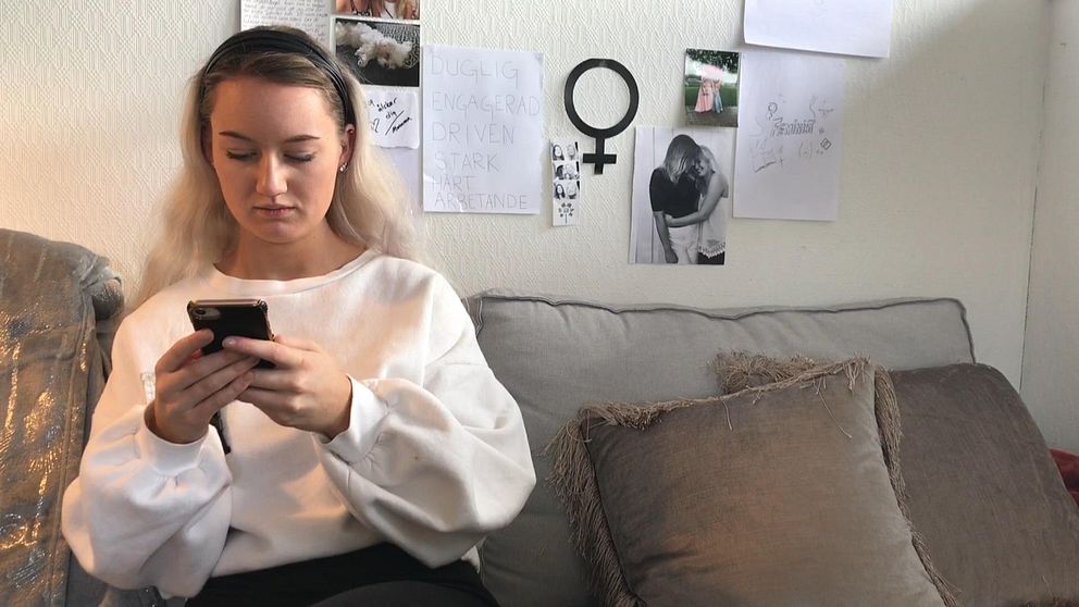 Lisa sitter i soffan med vit tröja hon använder mobilen, på väggen bakom hänger bilder.