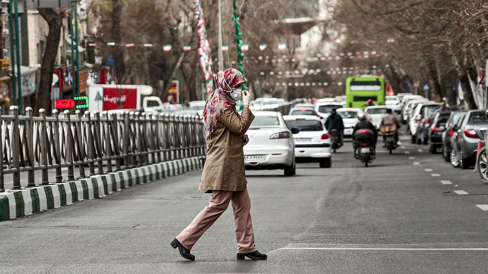 Bild på en kvinna i Iran som korsar en väg och har på sig ansiktmask och latexhandskar.