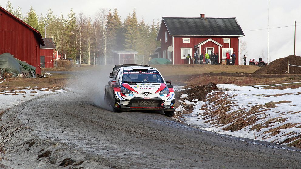 En rallybil på en grusväg med lite snö vid sidan. I bakgrunden syns lador och en röd stuga med vita knutar.