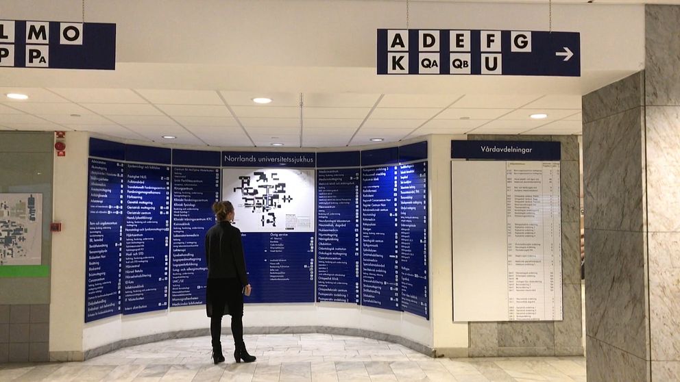 En kvinna i mörka kläder står och tittar på orienteringstavland i centralhallen på Norrlandsuniversitetsjukhus. I förgrunden syns skyltar och pilar som är upphängda från taket.