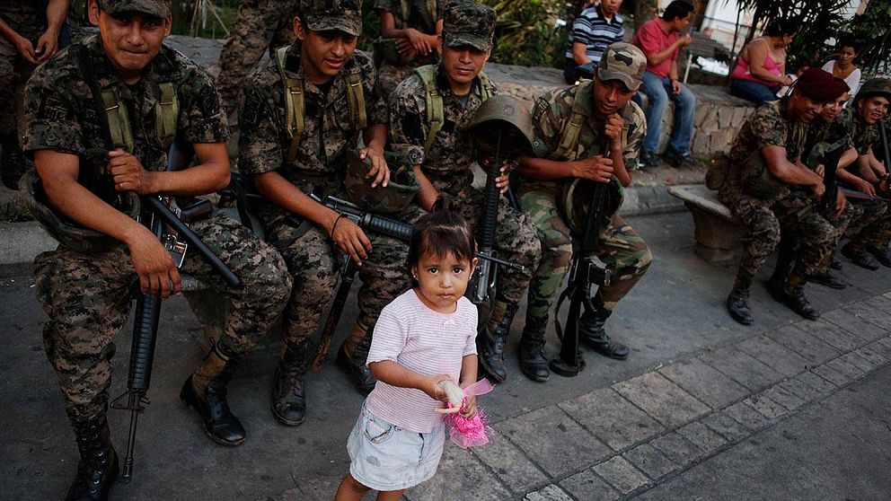 En liten flicka står bredvid honduranska militärer på rast på San Pedro Sulas centrala torg.