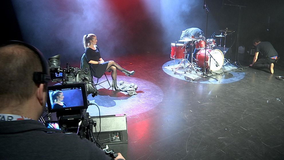 Emmy sitter på en stol på scenen, bredvid ett trumset. I förgrunden en tv-kamera som filmar henne.