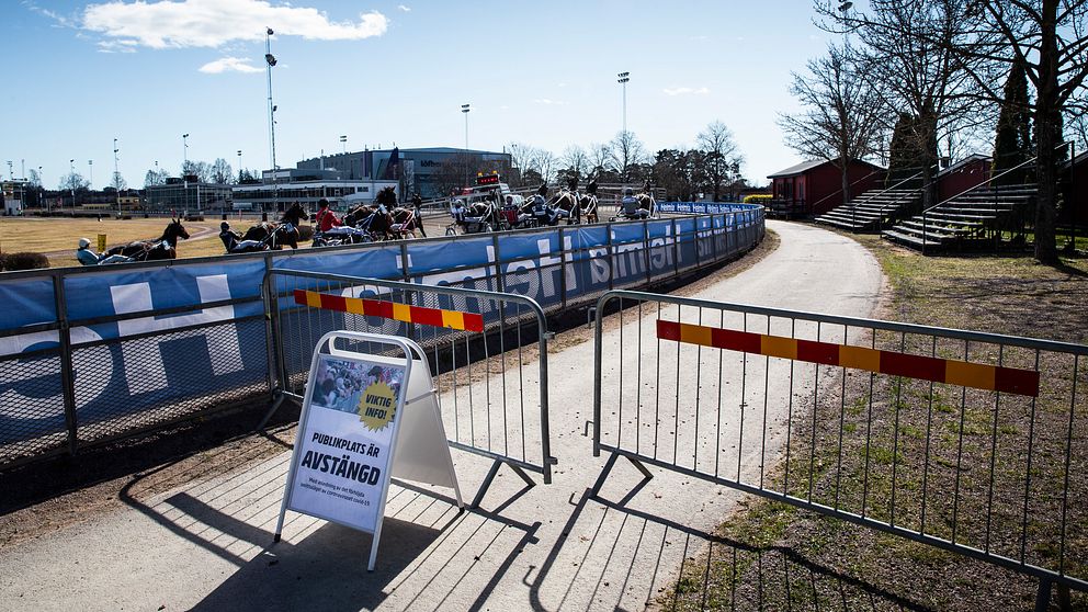 I förgrunden en skylt som visar att publikplatserna är avstängda, och i bakgrunden kör ekipagen i ett travlopp förbi på Färjestadstravet i Karlstad.