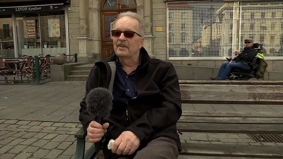 Thomas sitter på en parkbänk i centrala Linköping. I bakgrunden ser man en tom uteservering och en man i rullstol som passerar.