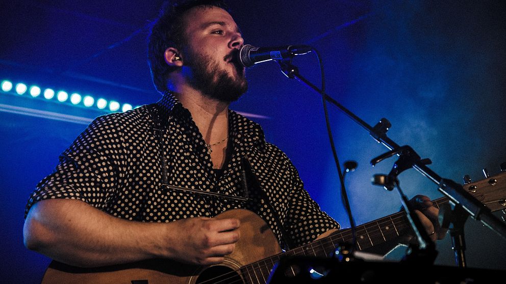 Trubaduren Christopher Edgar står på scenen i blått ljus och spelar gitarr och sjunger iklädd en svart skjorta med vita prickar.