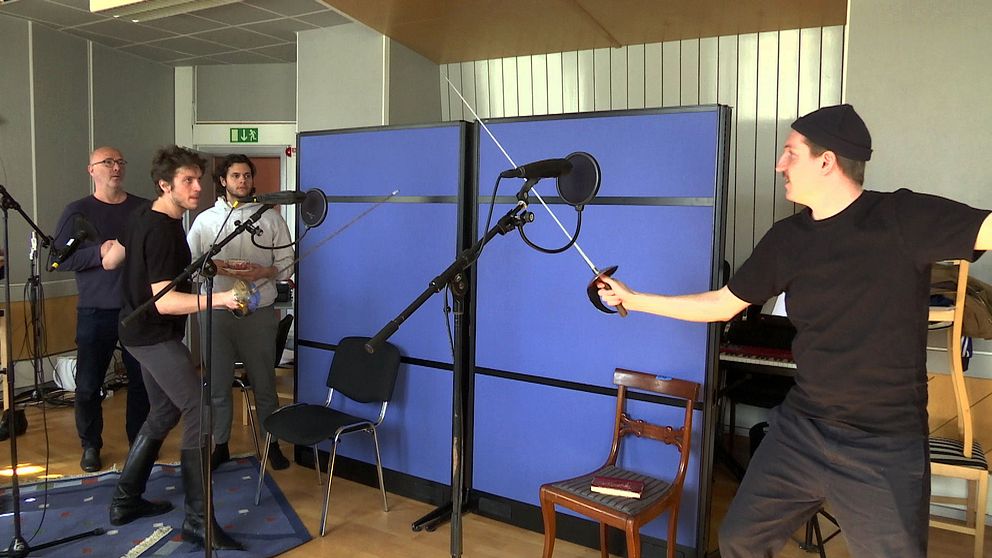 Två killar fäktas med glatt humör i en replokal. Flera mikrofoner syns uppställda och två andra män tittar intresserat på.