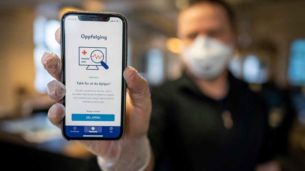 En man med ansiktsmask syns i bakgrunden. Han har plasthandskar på sig och håller upp en mobiltelefon där appen ”Smittestopp” syns.