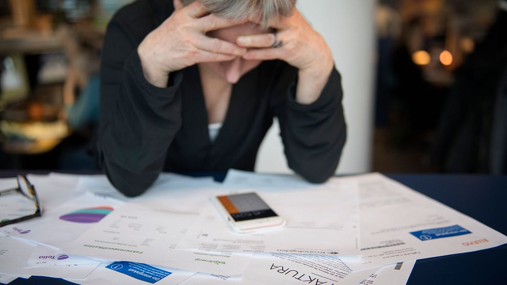 En kvinna sitter med huvudet lutat i händerna och tittar förtvivlat ner på bordet där det ligger en miniräknare ovanpå flera fakturor.