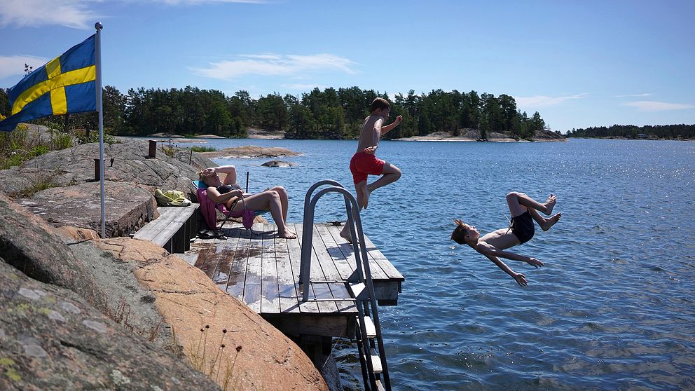 Två pojkar hoppar från en brygga. En kvinna solar sig på bryggan. Det är vackert väder, en svensk flagga vajar i vinden på en klippa intill bryggan.
