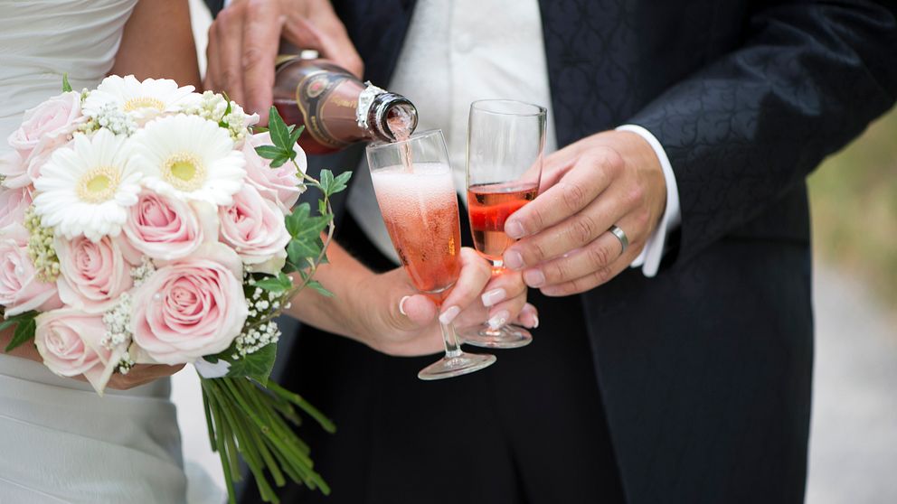 Närbild på brudpar, beskuren från axlarna och ned. Från vänster: bruden håller i en bukett av rosor och prästkragar medan brudgummen häller upp rosa champagne i hennes champagneglas.