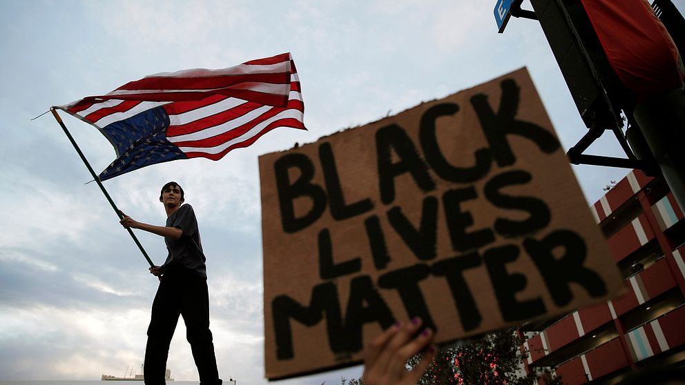 Ung man vajar USA:s flagga. En skylt med texten ”Black lives matter” syns också.