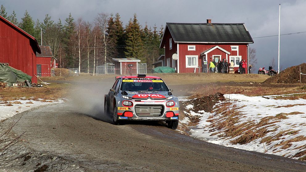 En rallybil på en grusig väg med en röd stuga samt lador i bakgrunden.