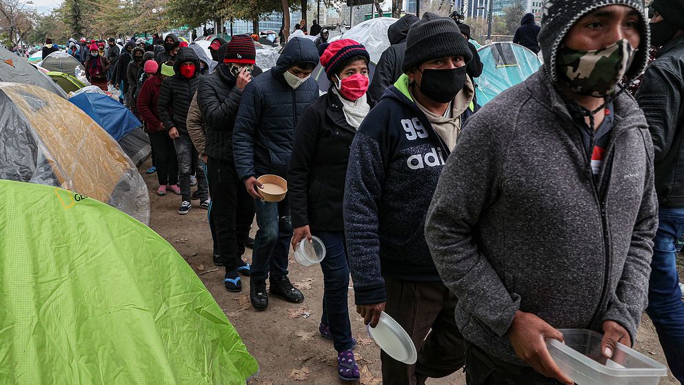 Fattiga migranter med munskydd och tomma matkärl i händerna köar utanför Bolivias ambassad i Santiago, Chile.