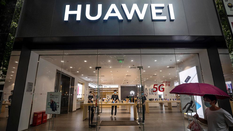 Bild tagen underifrån. En man med munskydd och paraply passerar skyltfönstret på en Huawei-butik.