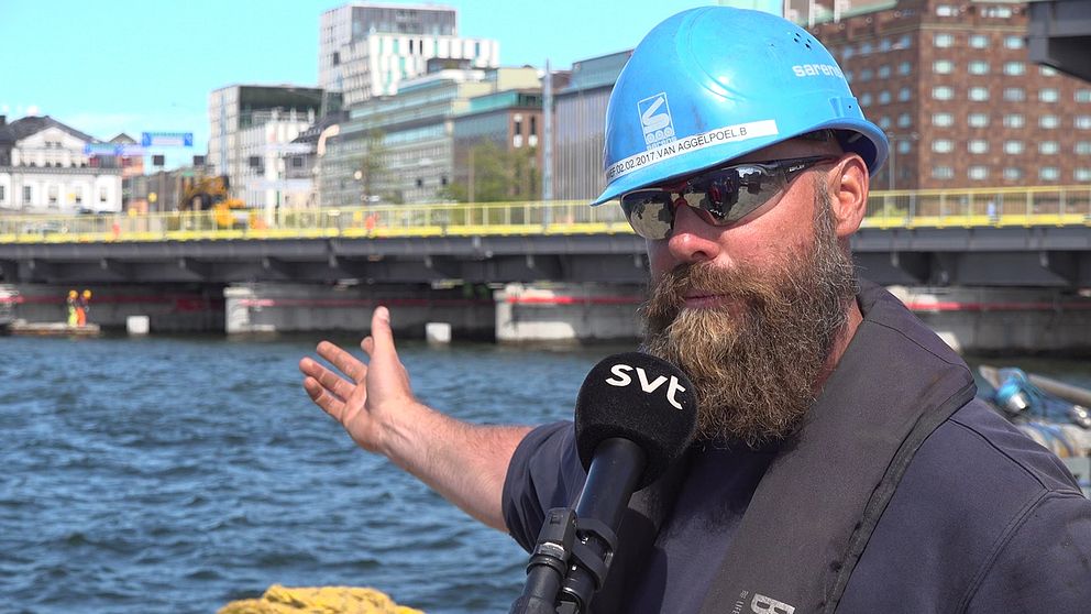 Man med blå bygghjälm står invid vattnet med centrala Stockholm som fond. Han pekar mot den bro som monteras bakom honom.