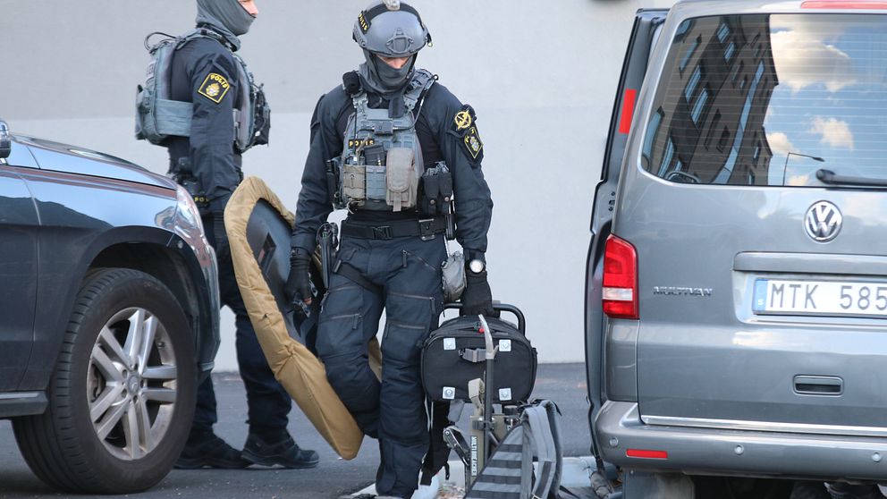 En polis med omfattande skyddsutrustning bär på en sköld. Intill honom står polisens piketbuss.
