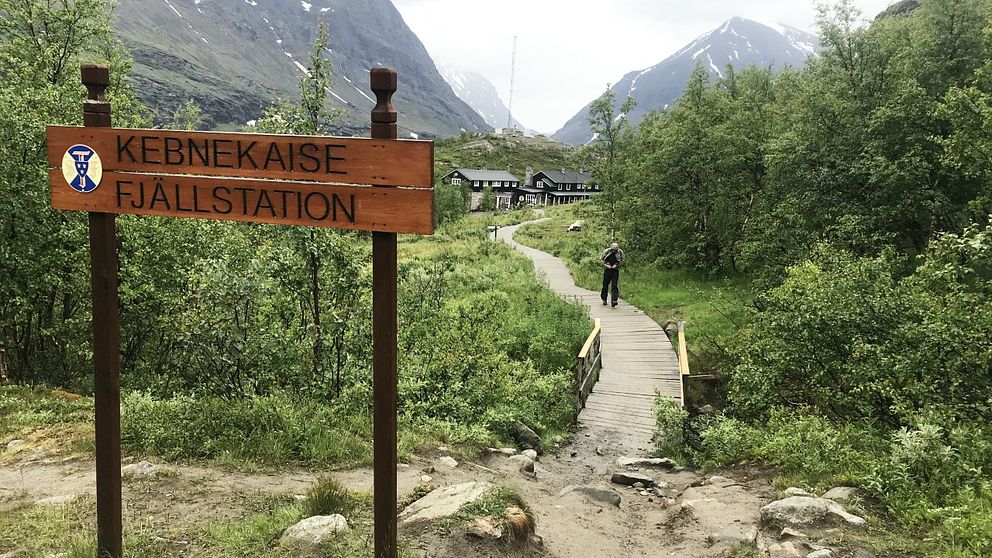 En träskylt där det står Kebnekaise fjällstation och en man som går på spångar. I bakgrunden syns fjäll och flera hus.