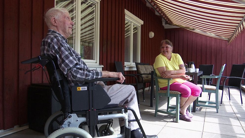 En äldre man i rullstol och en kvinna i färgglada kläder sitter och samtalar