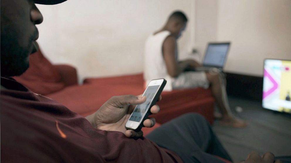 Två nätbedragare sitter i en lägenhet och kommunicerar med sina offer genom mobiltelefon och dator.