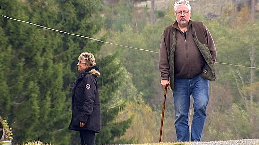 Leif GW går med käpp på grusväg, en äldre medelålders kvinna syns, skogsmark i bakgrunden
