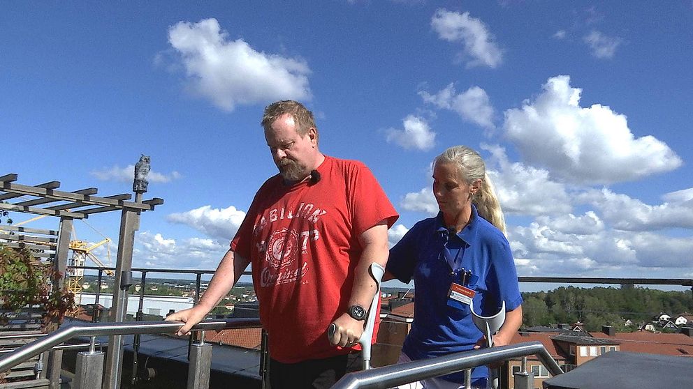 Johan står på en liten trappa uppe på en takterass med en molning himmel bakom. Han har kryckor och ansiktet är sammanbitet. Bredvid honom står fysioterapeuten Marina med en hand på hans rygg.